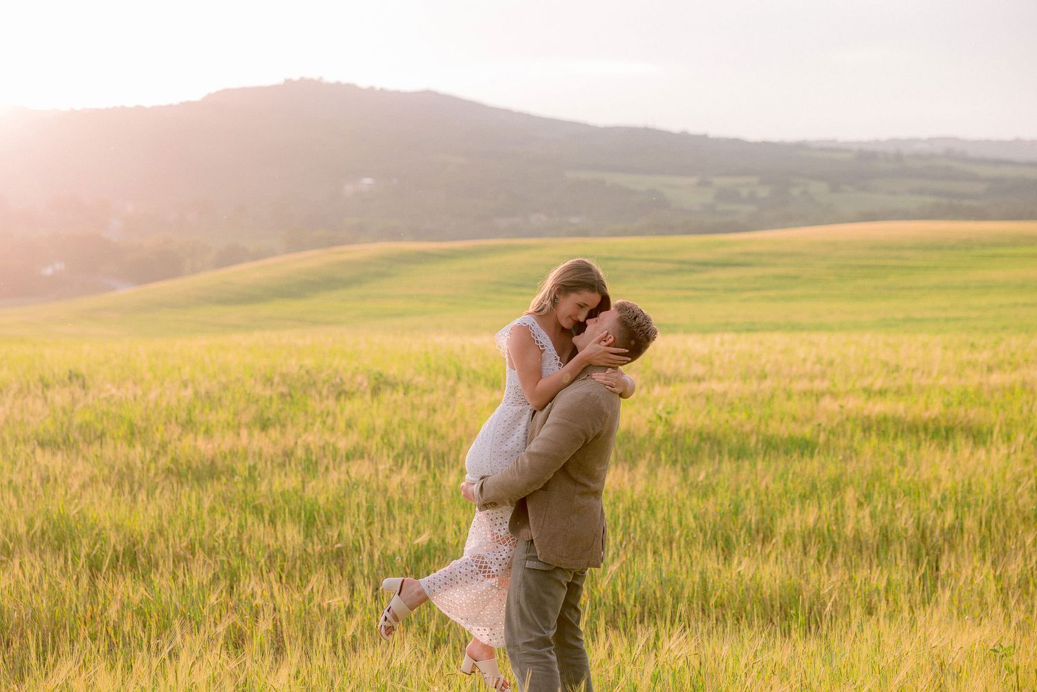 Two people share an intimate moment in a sun-drenched meadow with expansive countryside views.