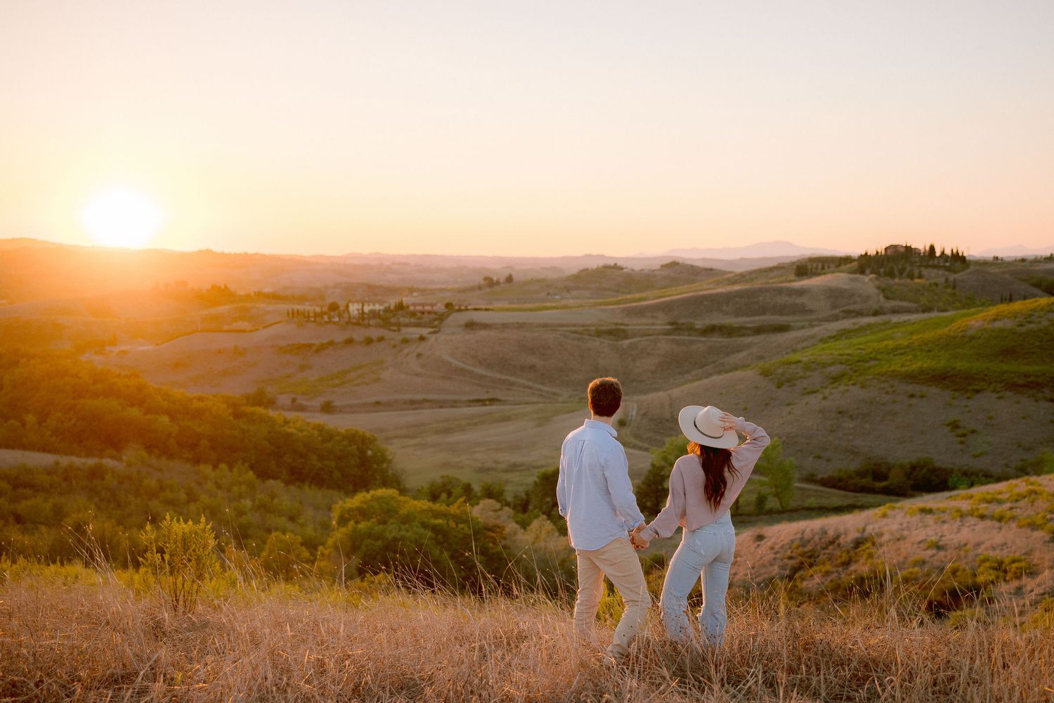 A couple stands together watching sunset over rolling hills in the countryside.