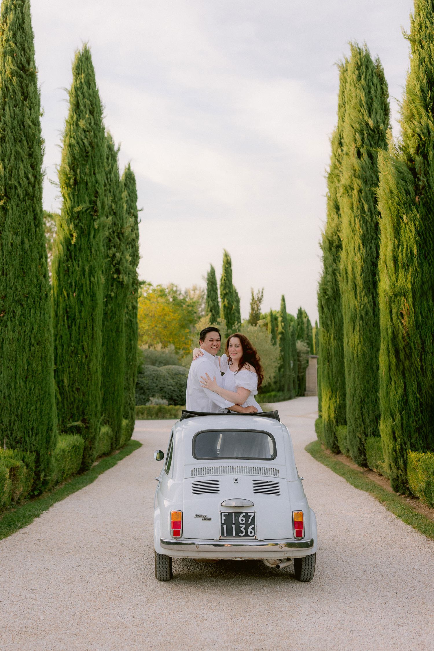 A vintage white Fiat car parked on a cypress-lined road in the Italian countryside.