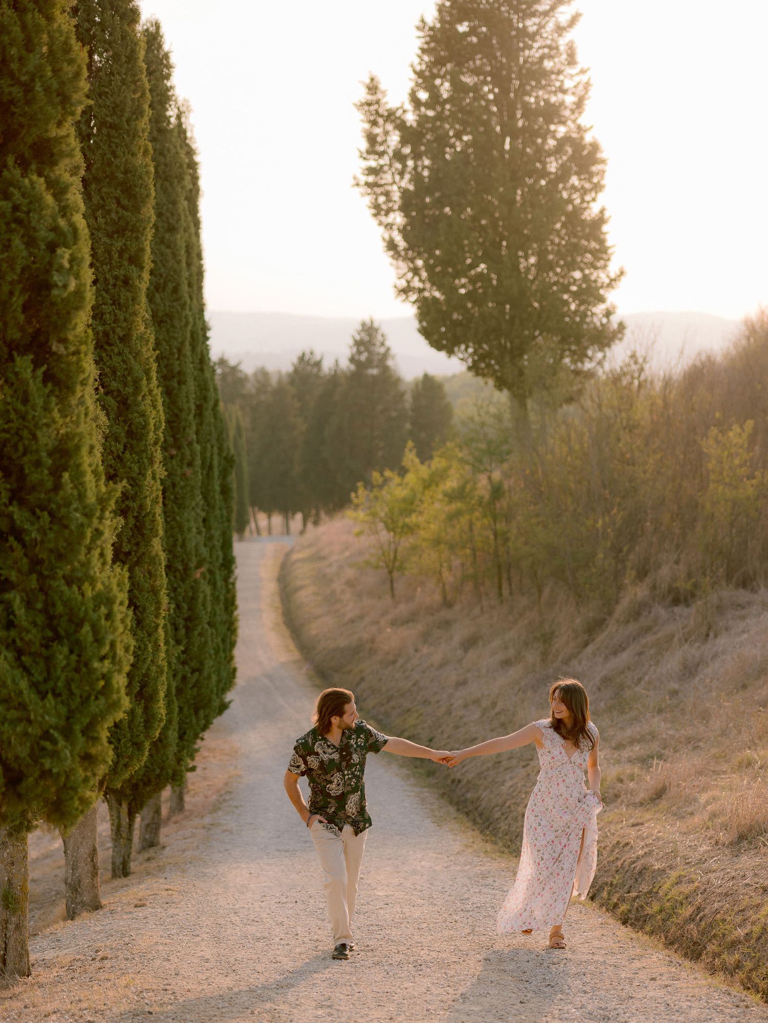 A couple walks hand in hand down a cypress tree-lined path in Tuscany.