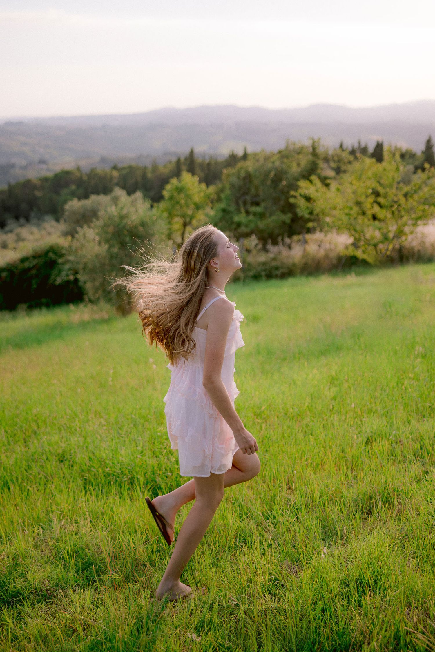 A person in a white dress twirls in a green meadow with mountains in the background.