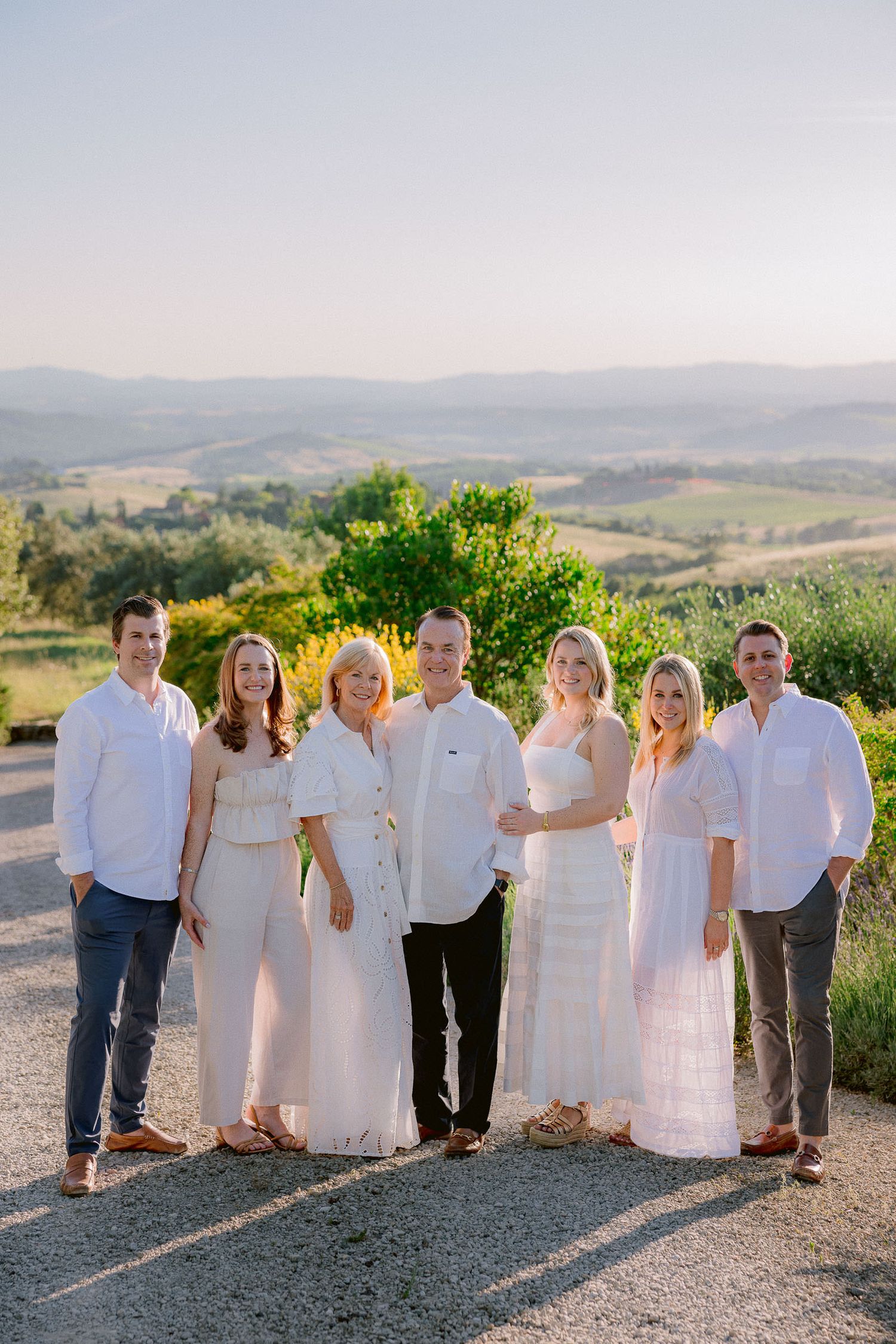 A group in white and cream colored outfits poses together on a gravel path with hills in the background.