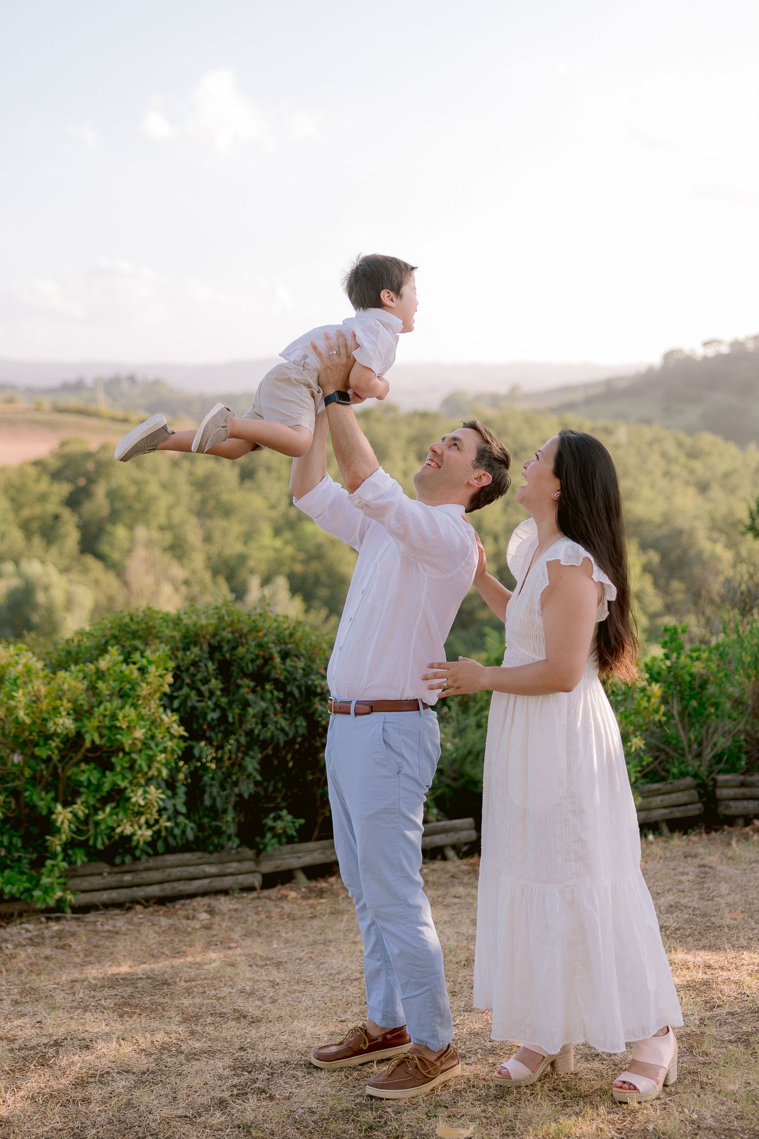 A couple playfully lifts a small child against a scenic vineyard backdrop at sunset.