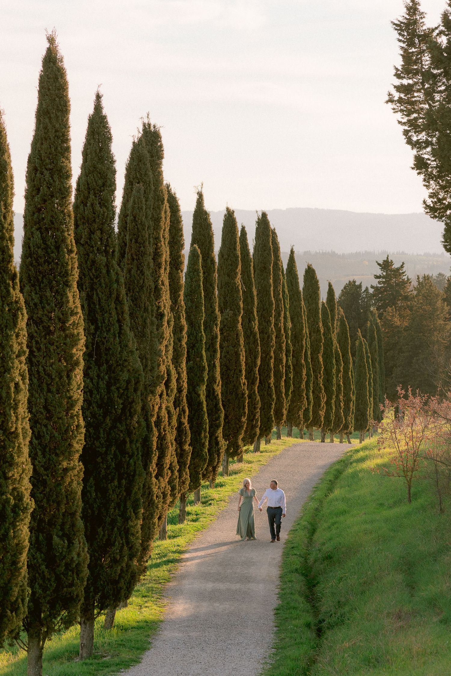 Two figures strolling down a cypress tree-lined path in the Italian countryside.