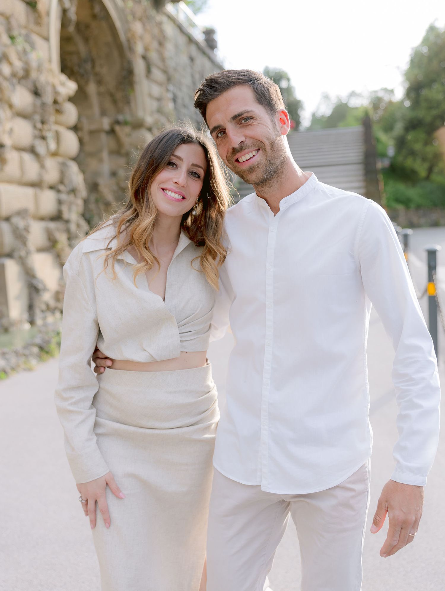 Couple in matching white outfits posing against a rustic stone wall.