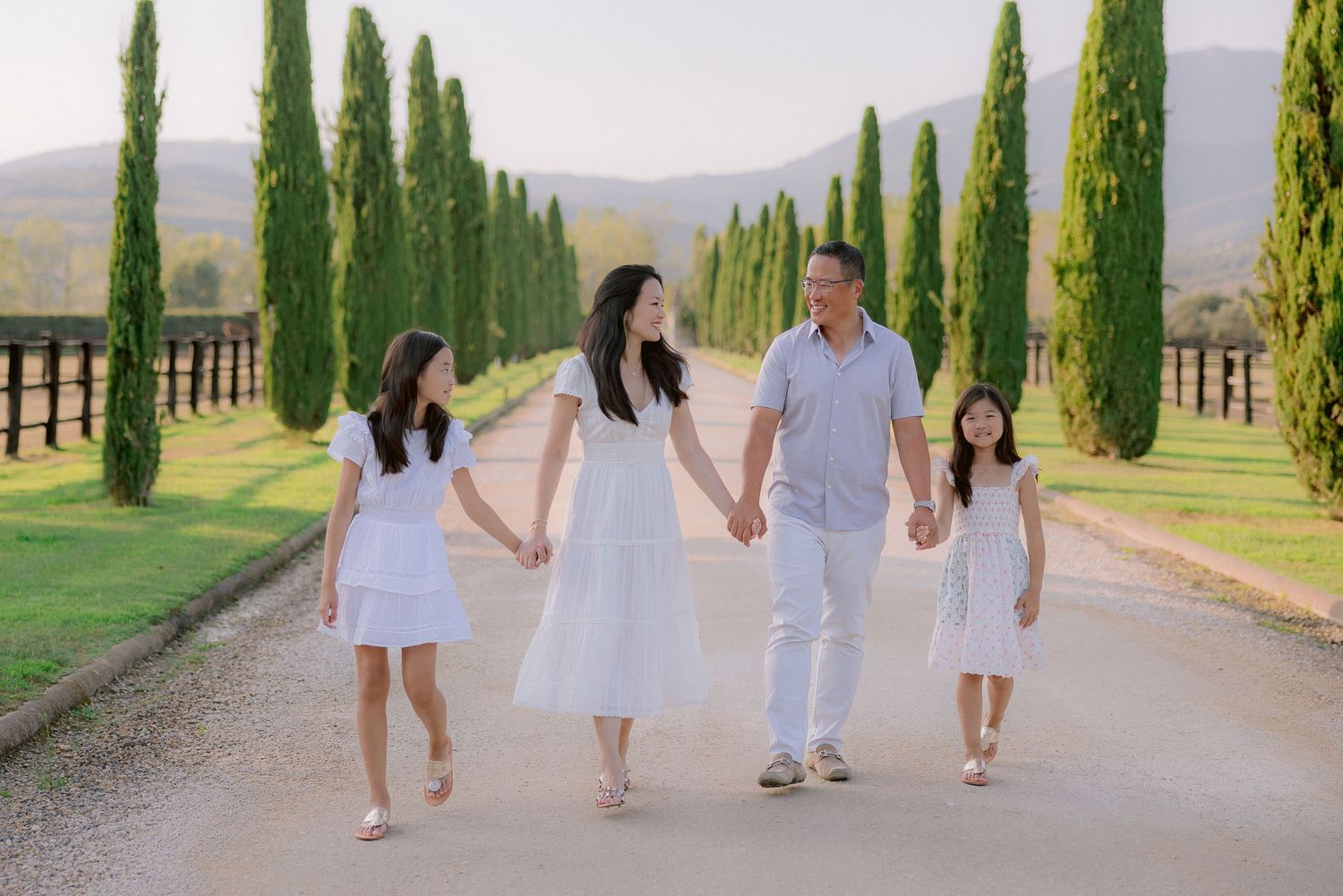 Family walking along cypress tree-lined path in coordinated white outfits.