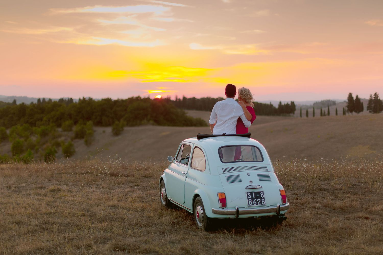Vintage light blue Fiat car against dramatic sunset sky in Tuscan countryside.