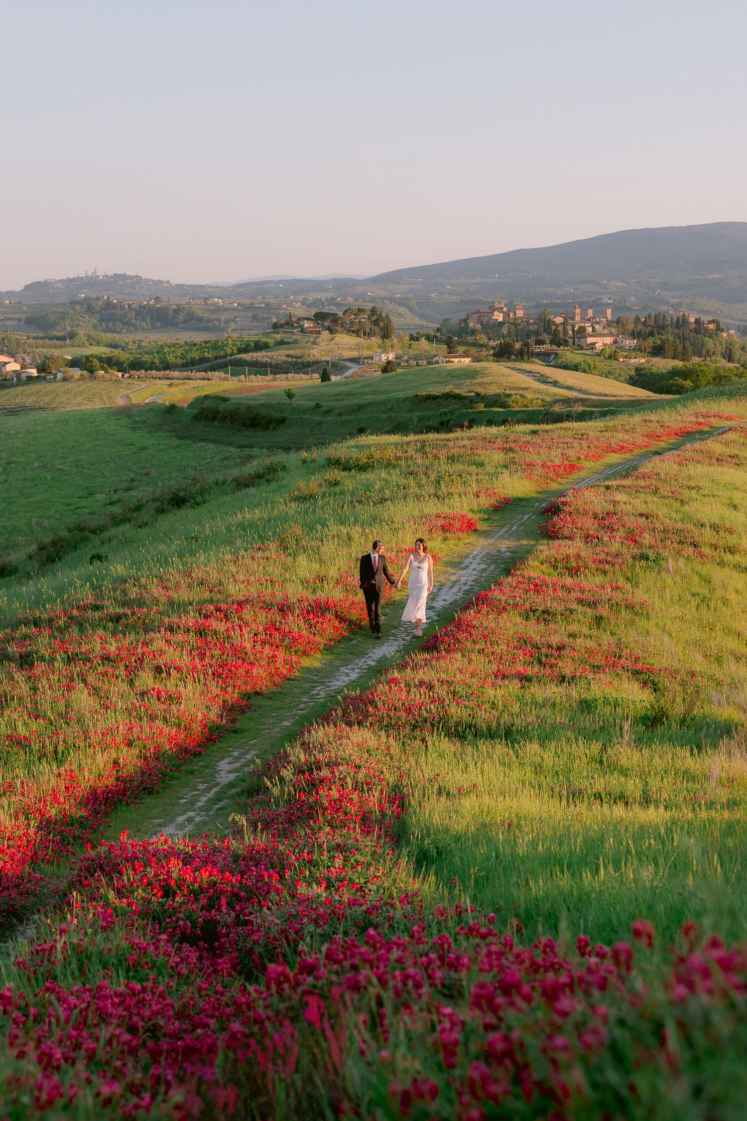 Two figures walk along a path through vibrant red wildflowers in rolling Tuscan hills at sunset.