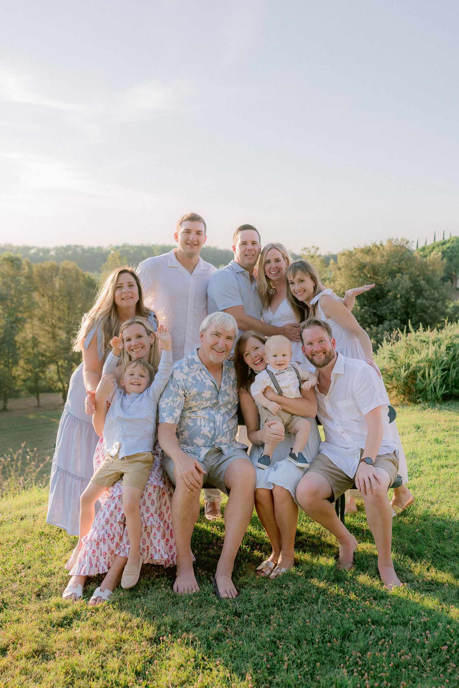 A multi-generational family group poses together on a grassy hillside wearing coordinated light-colored outfits.