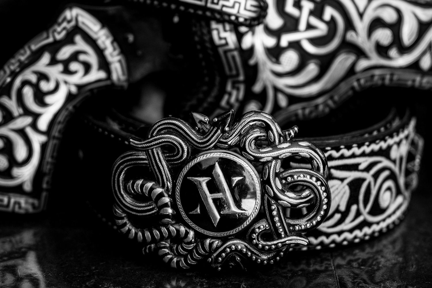 Black and white photo of an ornate silver belt buckle with monogram HH on a decorative leather belt.