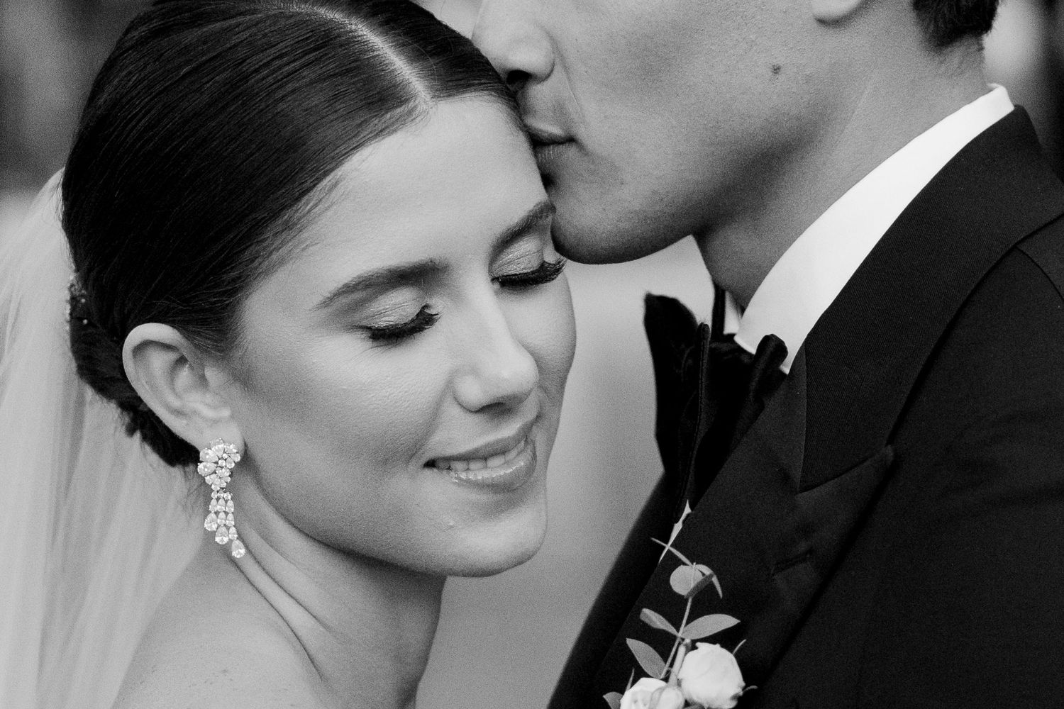 Black and white wedding photo of bride smiling as groom kisses her cheek, both elegantly dressed.
