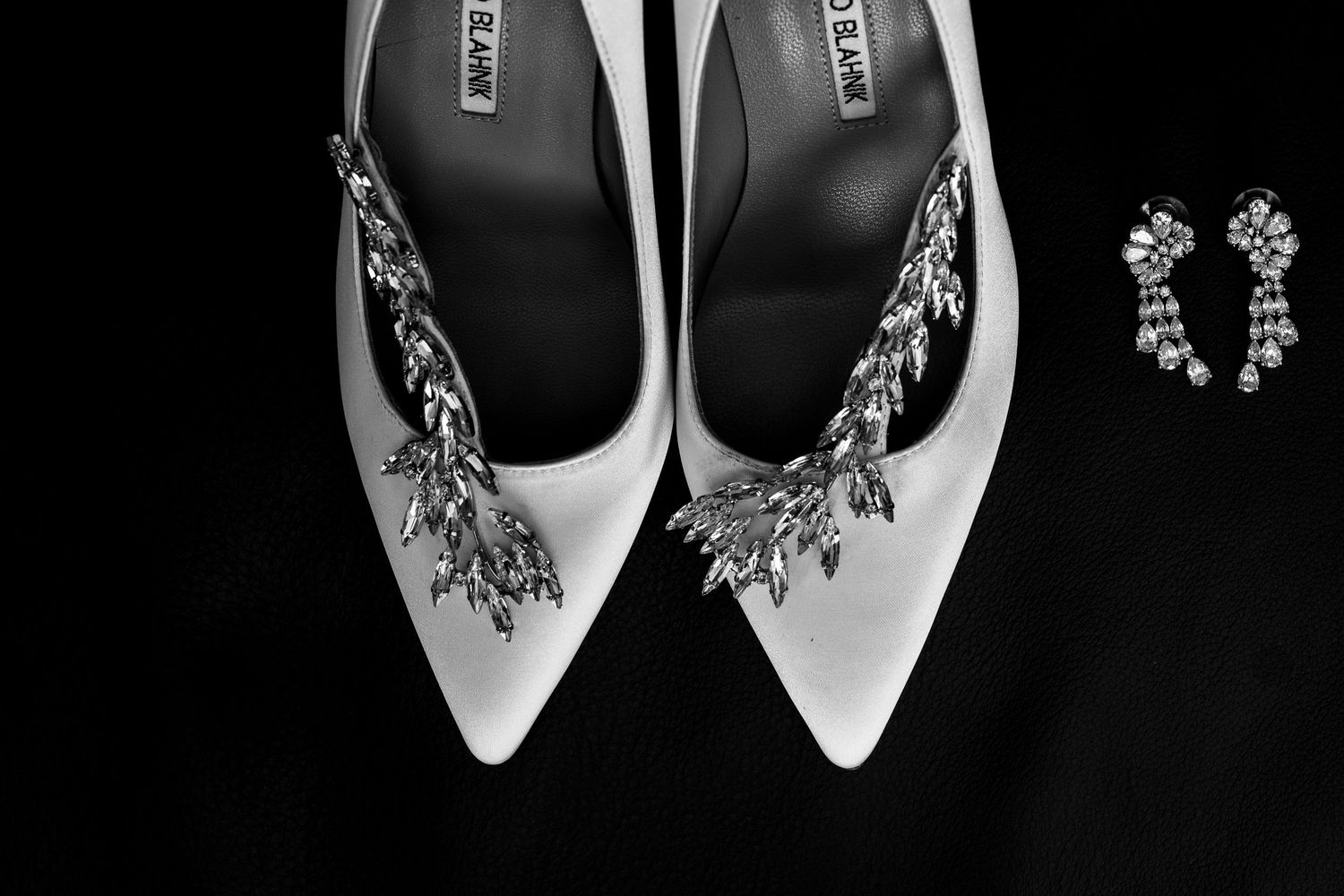 Black and white photo of white bridal heels with crystal embellishments beside pearl and diamond drop earrings.