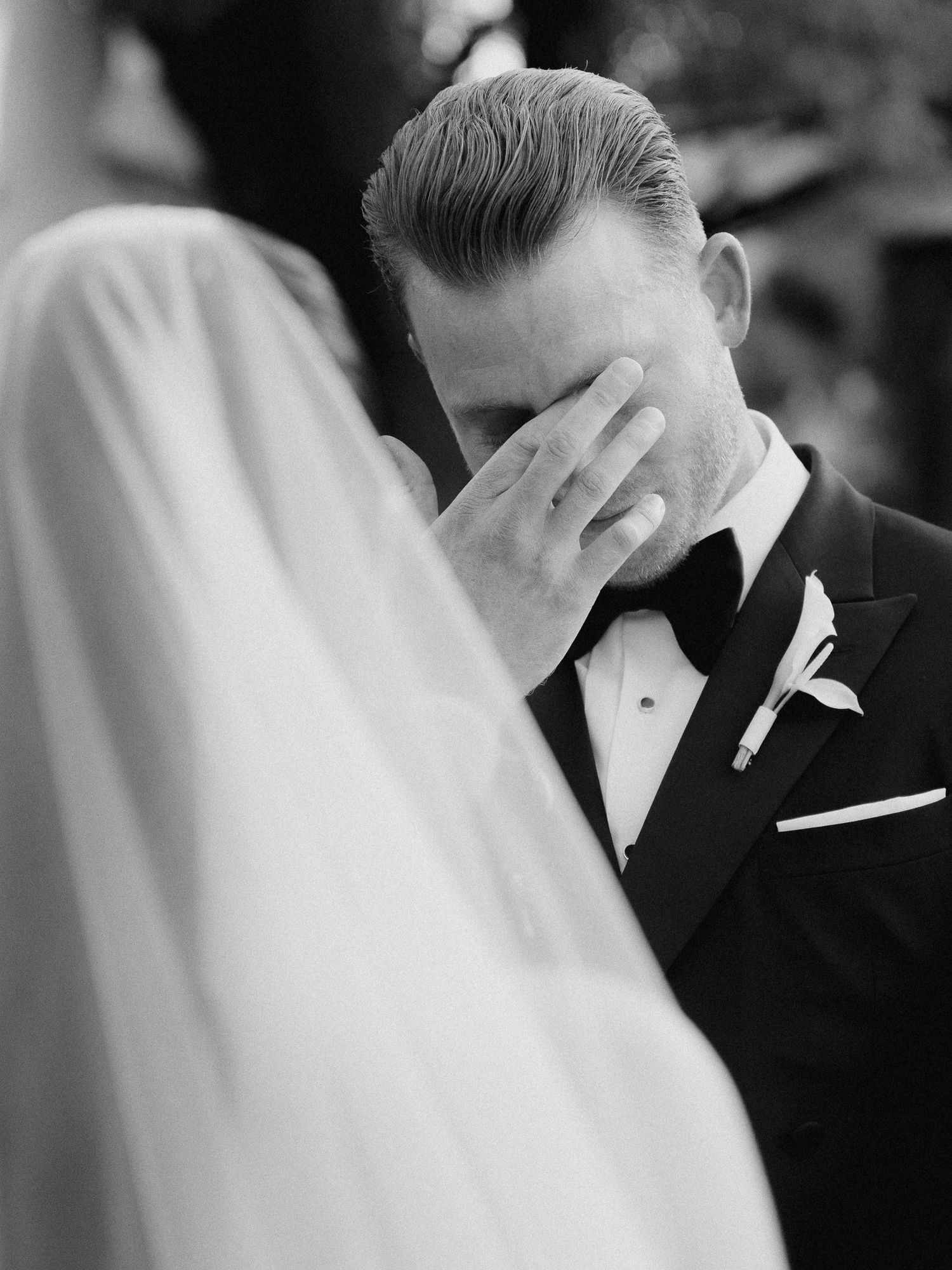 Emotional groom in tuxedo covering his face with tears during wedding ceremony, black and white photo.