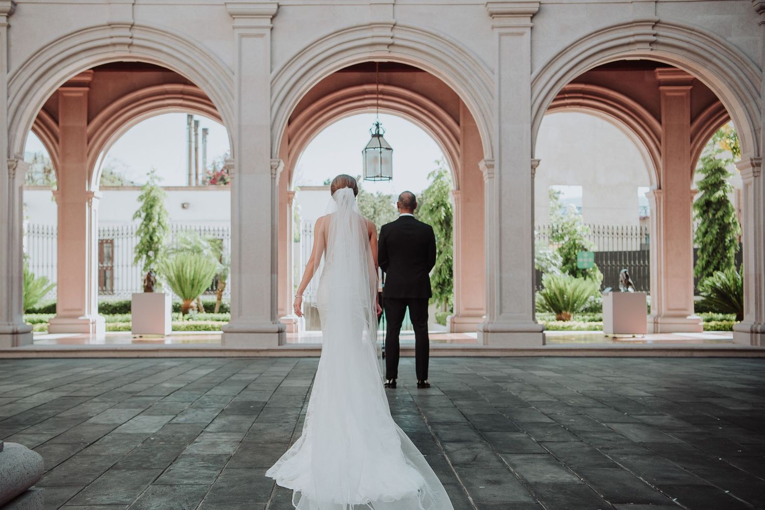 Bride and groom stand hand in hand beneath elegant pink arched colonnade, with lush garden and tower in background.