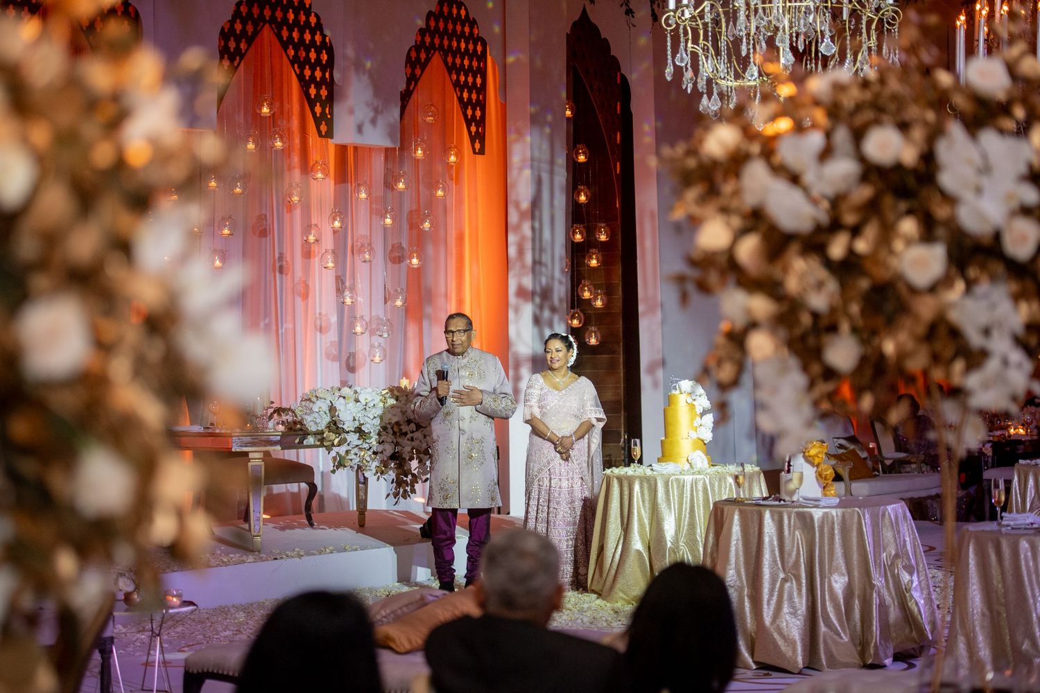 Elegant wedding reception with couple standing before floral decor, warm lighting, and chandelier in ornate venue.