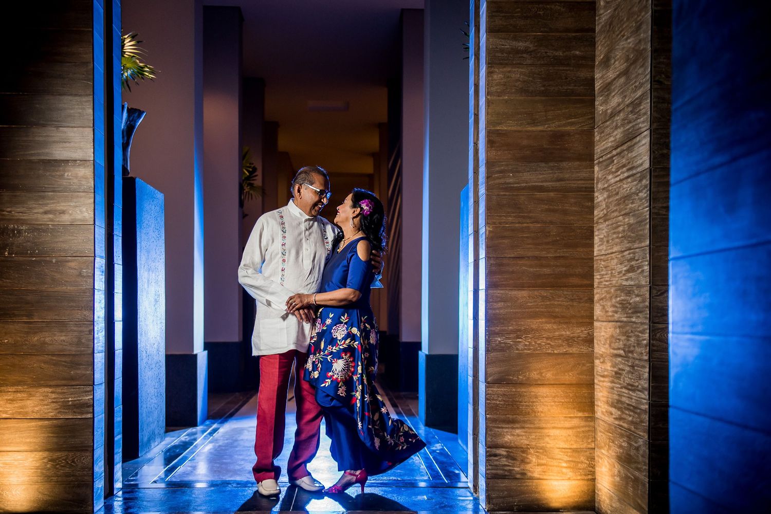 Couple in vibrant traditional attire share a romantic moment in a beautifully lit corridor during an Indian wedding celebration.