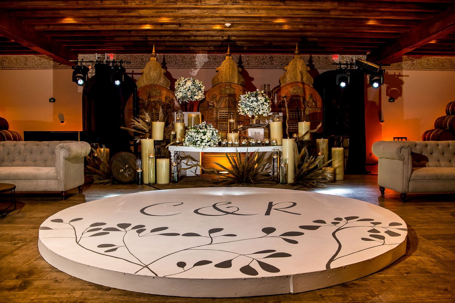 Elegant wedding reception with monogrammed dance floor, floral centerpieces, candles, and warm amber lighting in a rustic venue.