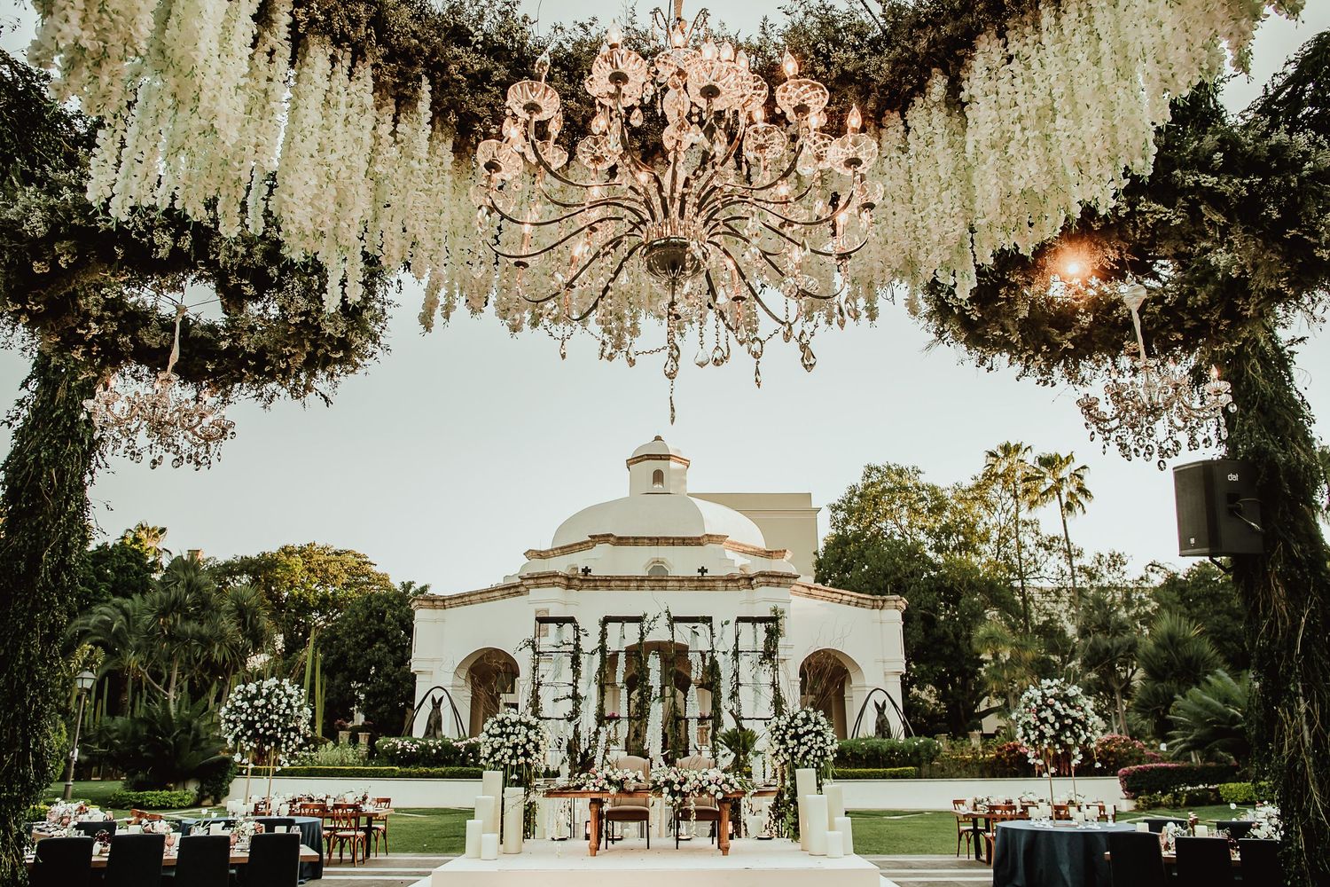 Elegant outdoor wedding venue with crystal chandelier, lush floral arch, and grand white colonial building backdrop.