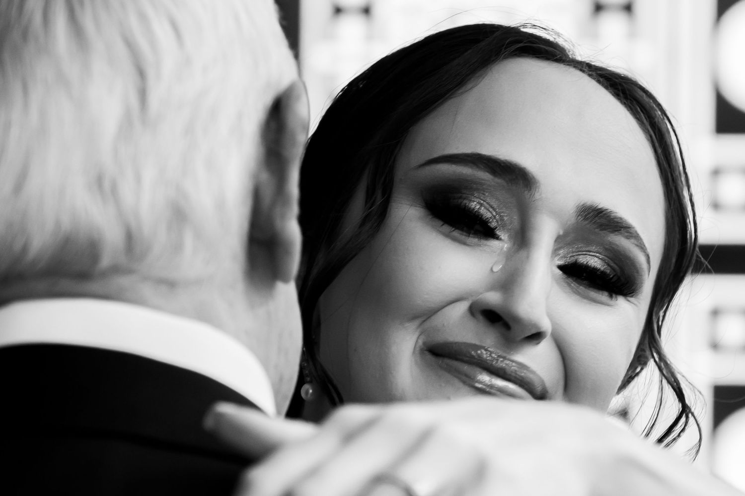 A black and white close-up portrait of a smiling woman with dark hair and elegant makeup during a tender moment.