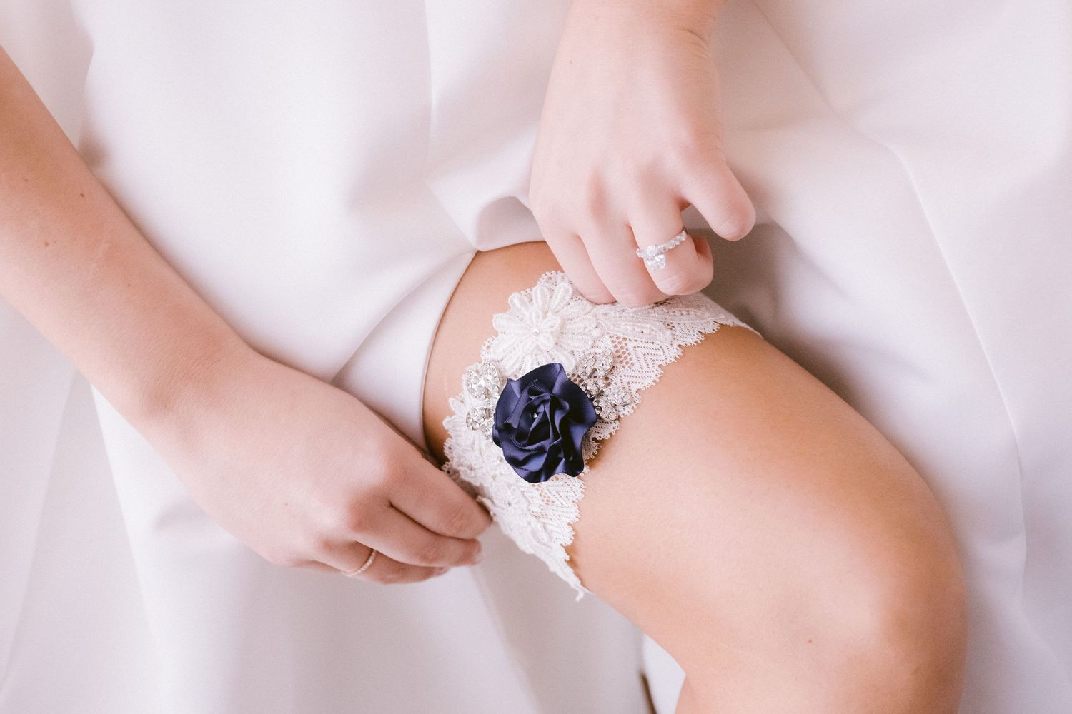 Hands adjusting a navy blue and lace wedding garter on white fabric.