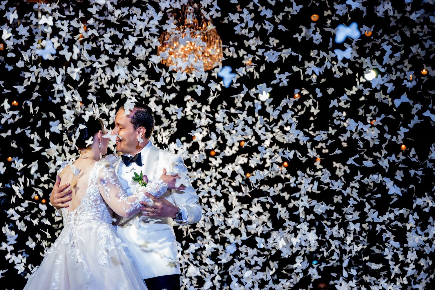 A romantic wedding dance moment surrounded by falling white confetti and chandelier lighting.