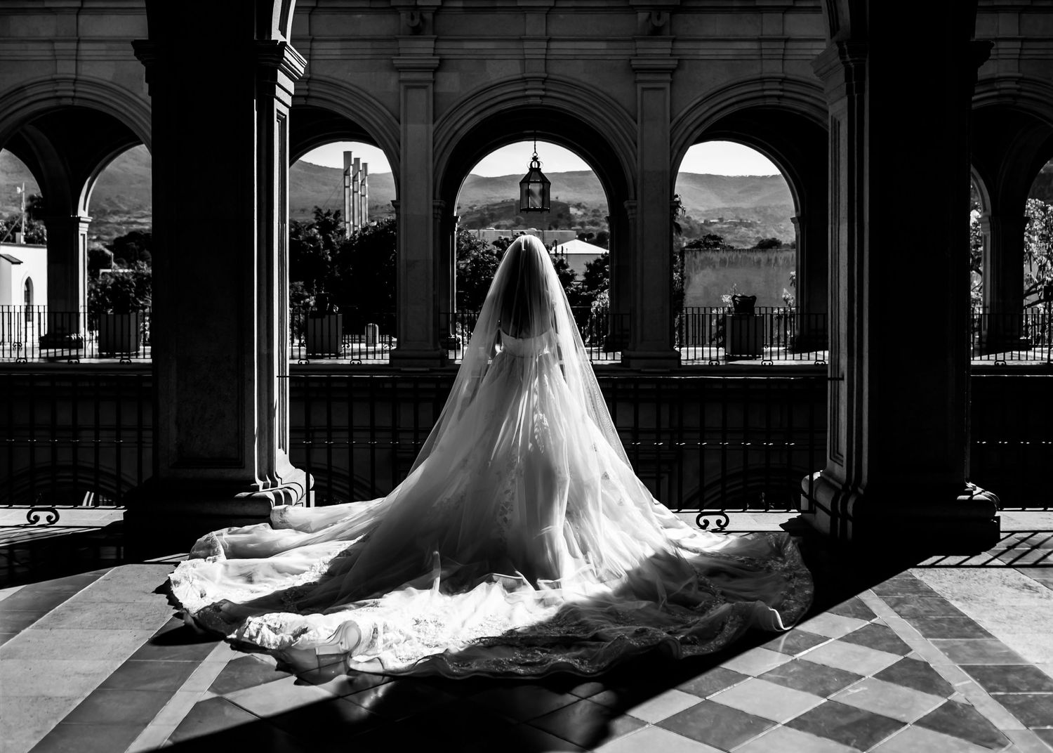 Black and white photo of a bride in a flowing gown and veil standing between elegant arched columns.
