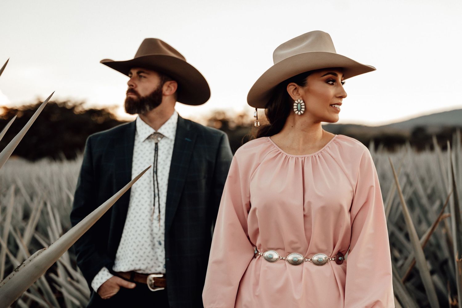 Stylish couple in western hats posing in agave field, woman in pink dress.