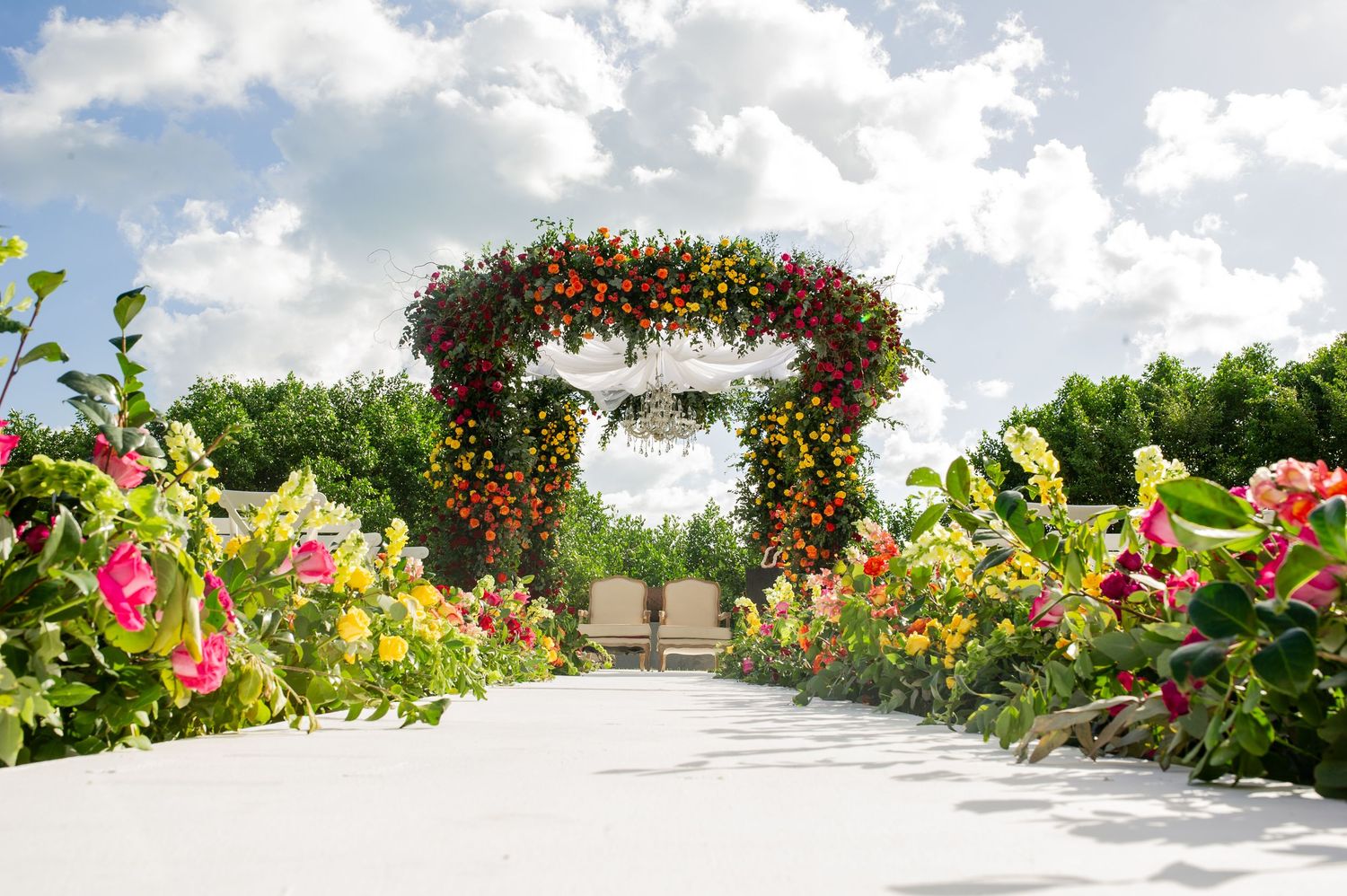 Colorful floral arch wedding ceremony backdrop with vibrant blooms lining a white pathway under a cloudy sky.