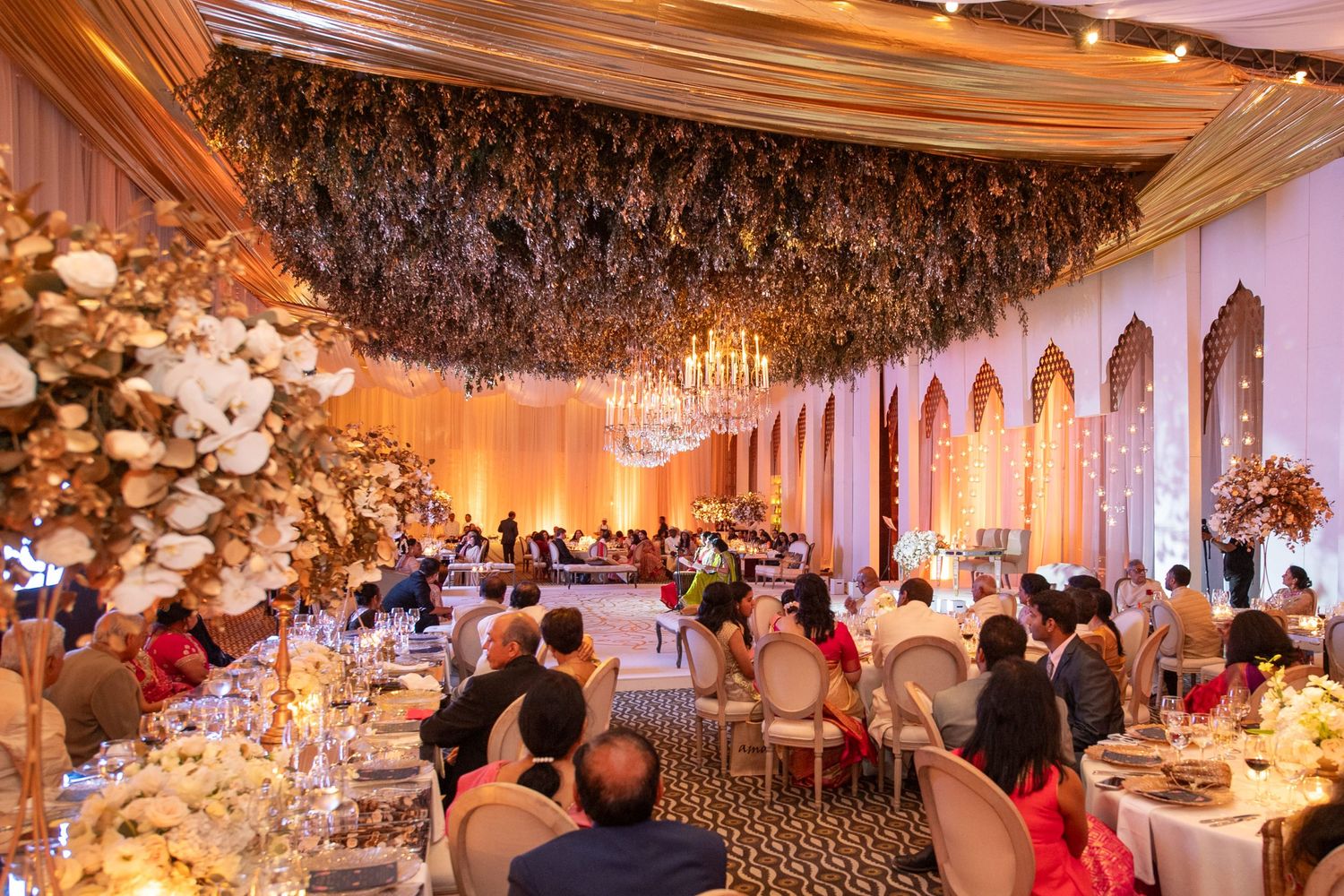 Elegant wedding reception hall with floral ceiling installation, chandelier, gold decor, and guests seated at decorated tables.