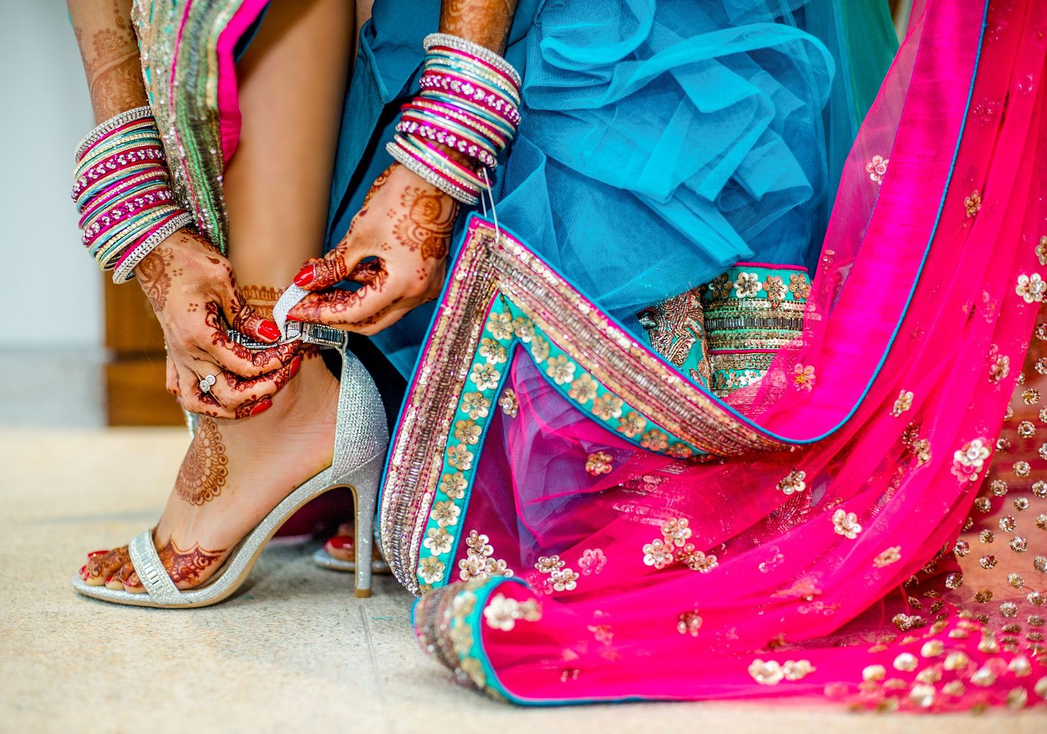 Bride in vibrant pink and teal lehenga with mehndi hands adjusting silver heels, colorful bangles adorning her wrists.