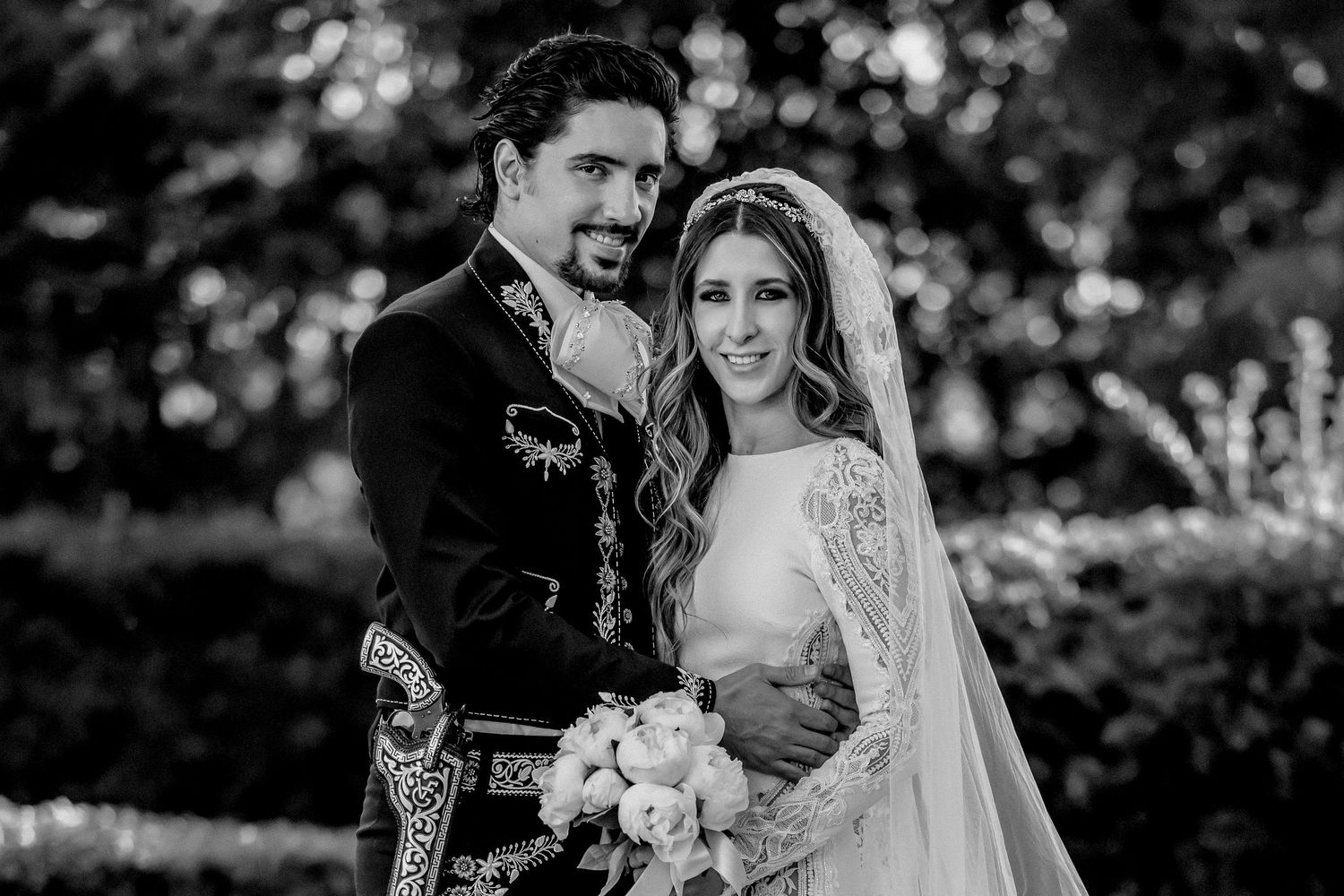 Black and white wedding photo of elegant couple, bride in lace veil holding bouquet, groom in embroidered suit.