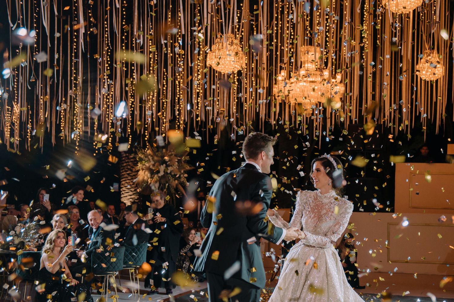 Bride and groom dancing at elegant wedding reception with golden confetti and warm string lights.