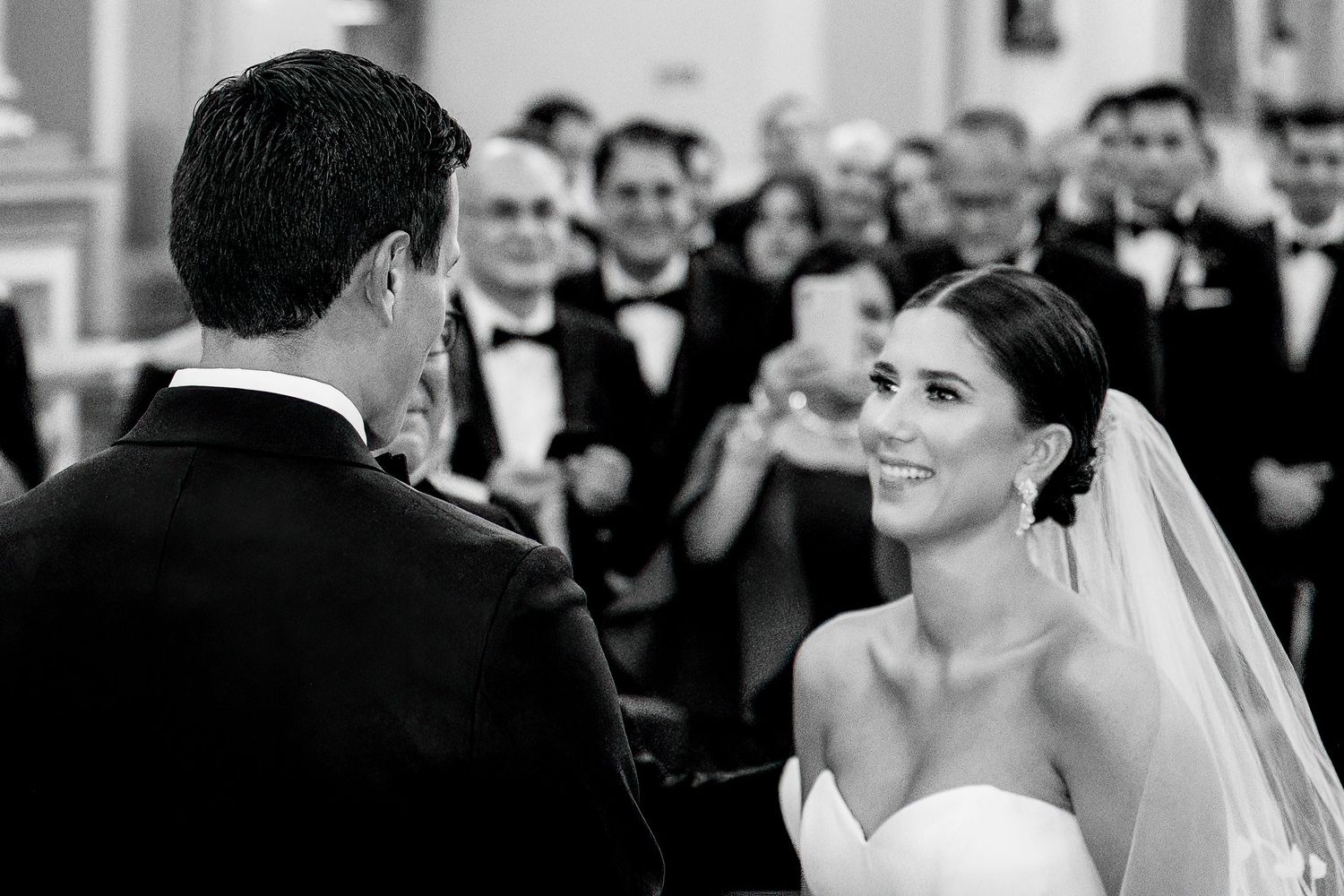 Black and white wedding ceremony photo of smiling bride in veil facing groom at altar with guests in background.