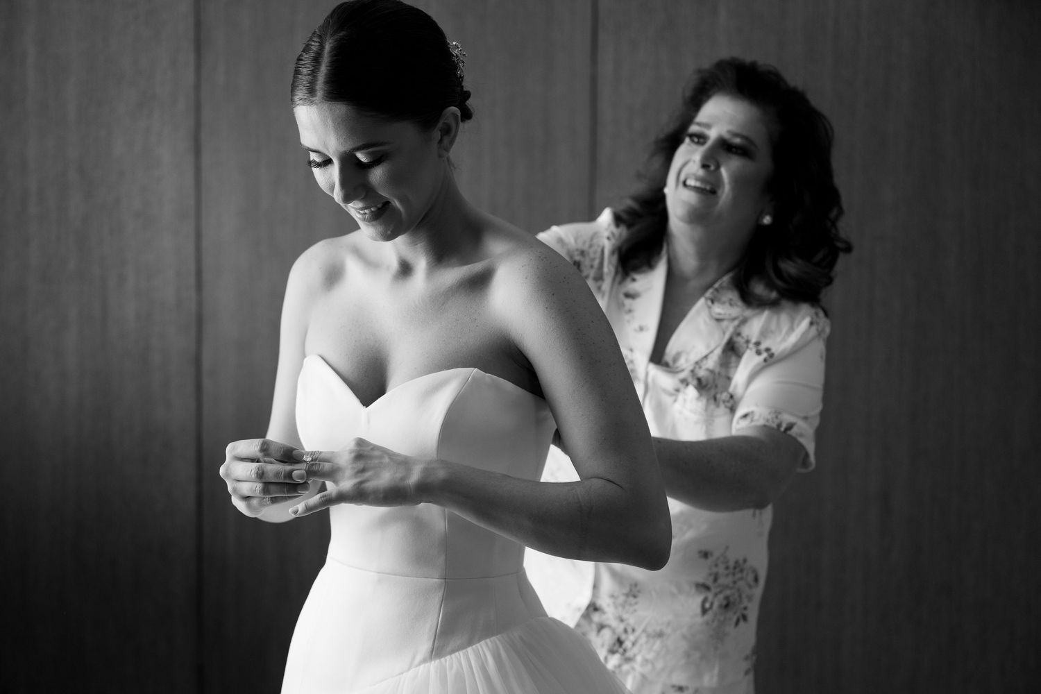 Bride in strapless white gown being helped by woman in floral dress, black and white wedding preparation photo.
