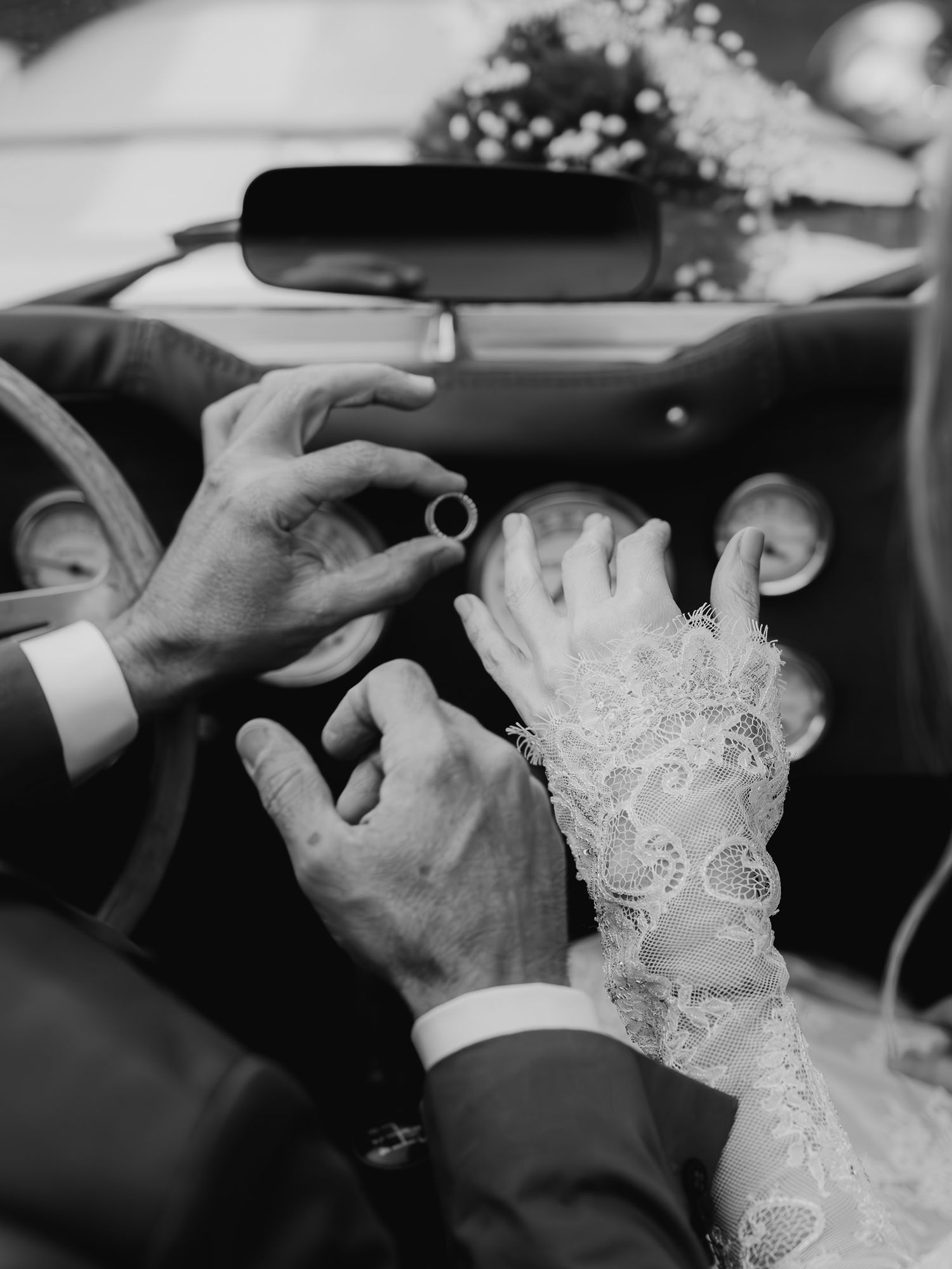 Black and white photo of groom placing wedding ring on bride's lace-gloved hand inside a decorated car.