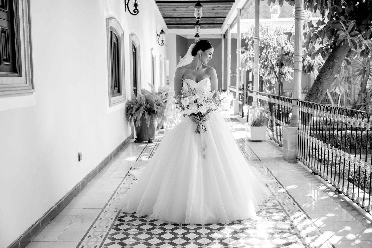 Bride in a white ball gown holding a bouquet poses on a tiled corridor in a black and white photo.