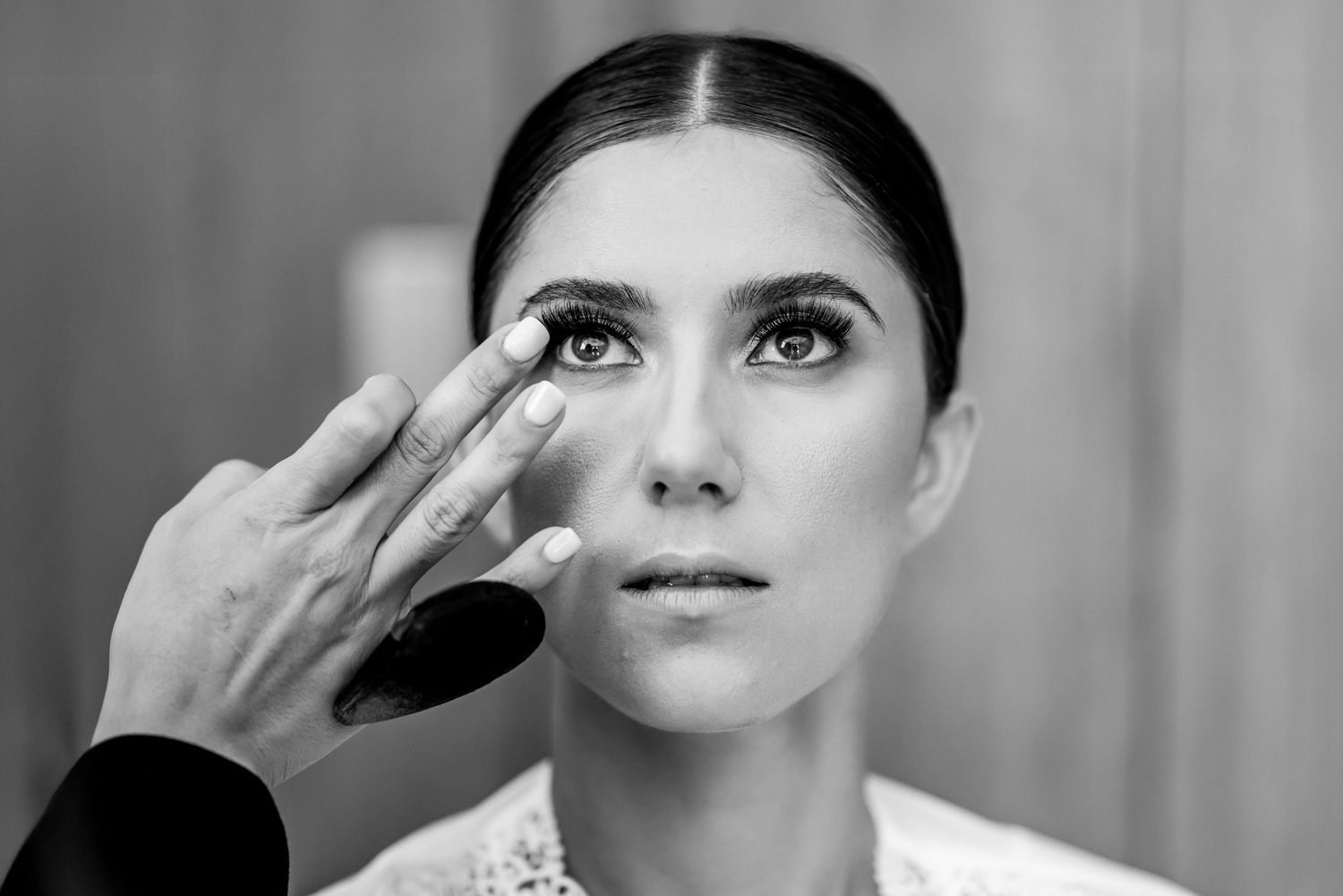 Black and white photo of a woman having eye makeup applied by a makeup artist before a wedding.
