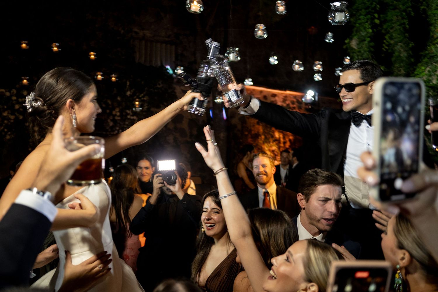 Guests celebrate at an elegant outdoor evening event, raising drinks and phones as a drone hovers above the crowd.