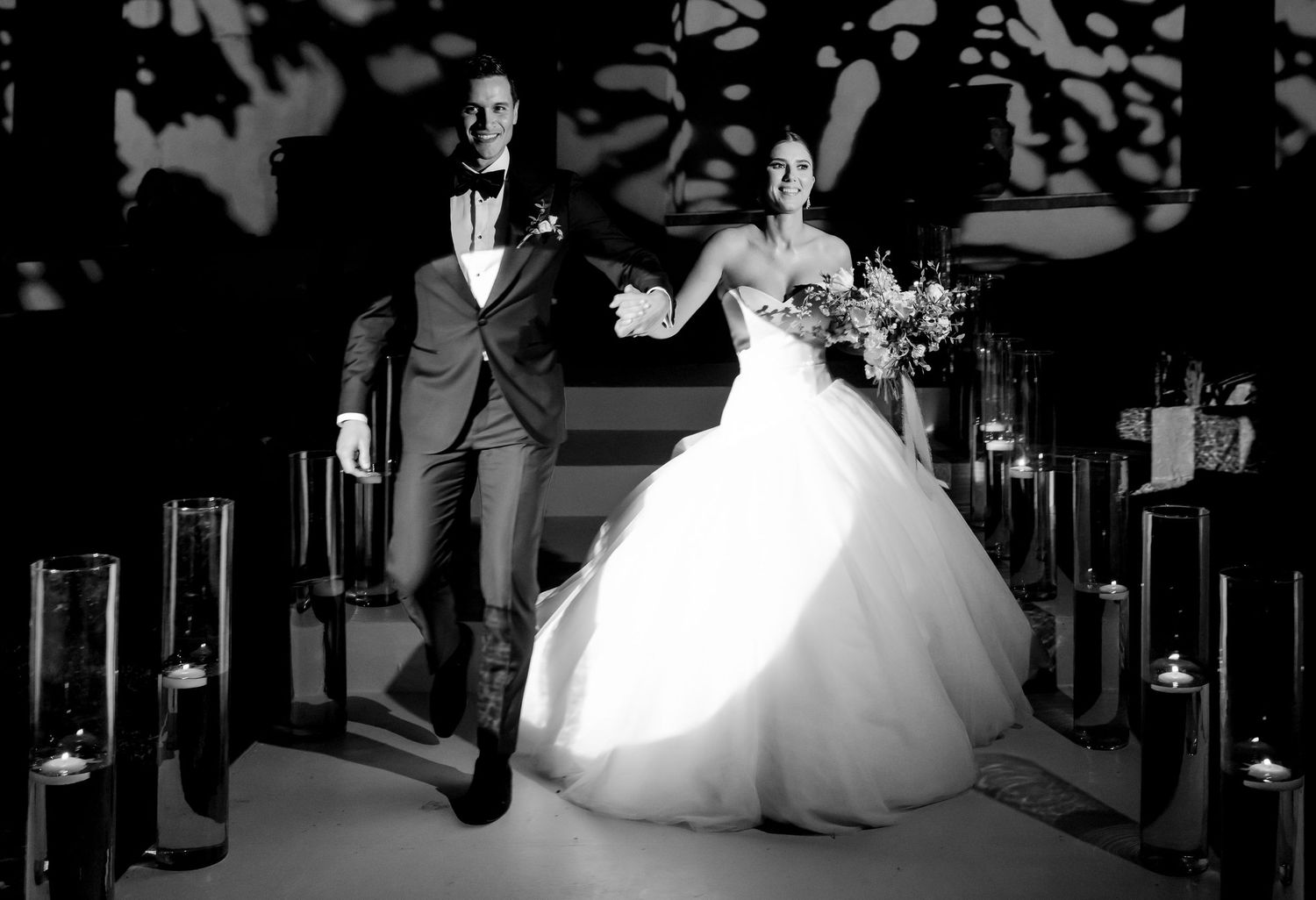 Bride and groom walk down candlelit aisle in stunning black and white wedding photo.
