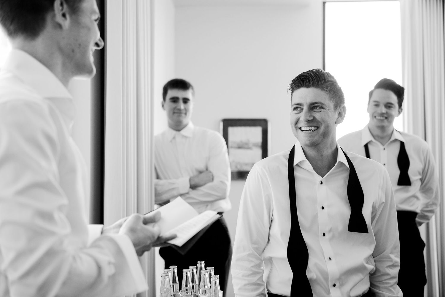 Groomsmen in white shirts and black ties laughing together while getting ready for a wedding.