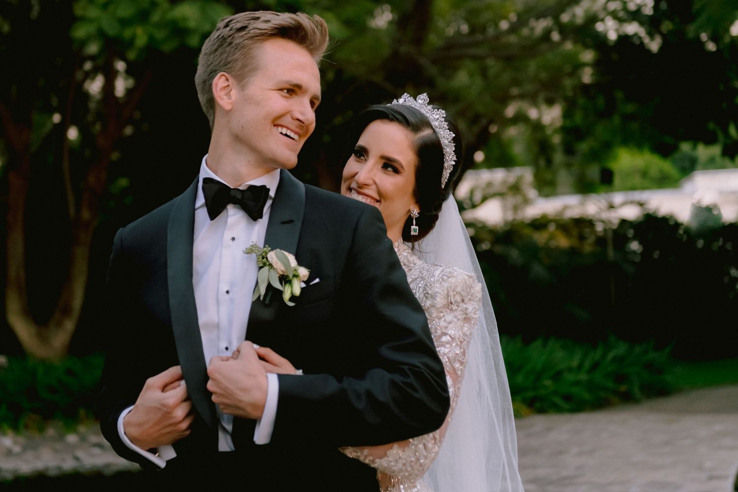 Elegant bride and groom posing outdoors on their wedding day, groom in black tuxedo, bride in lace gown with tiara.