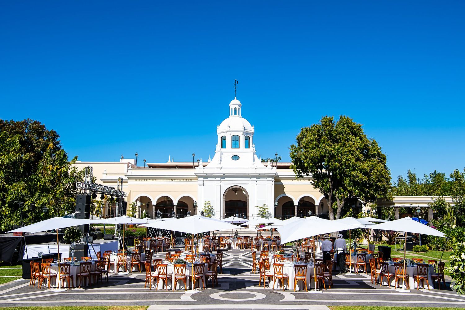 Elegant outdoor dining terrace with white umbrellas in front of a Spanish colonial building under a clear blue sky.