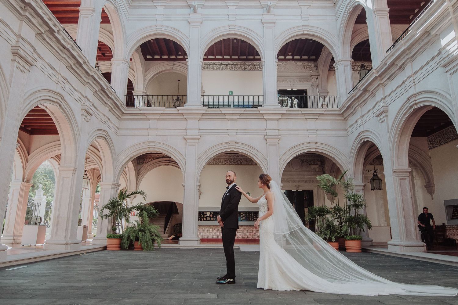 Bride and groom share a romantic first look in an elegant white arched courtyard with lush tropical plants.
