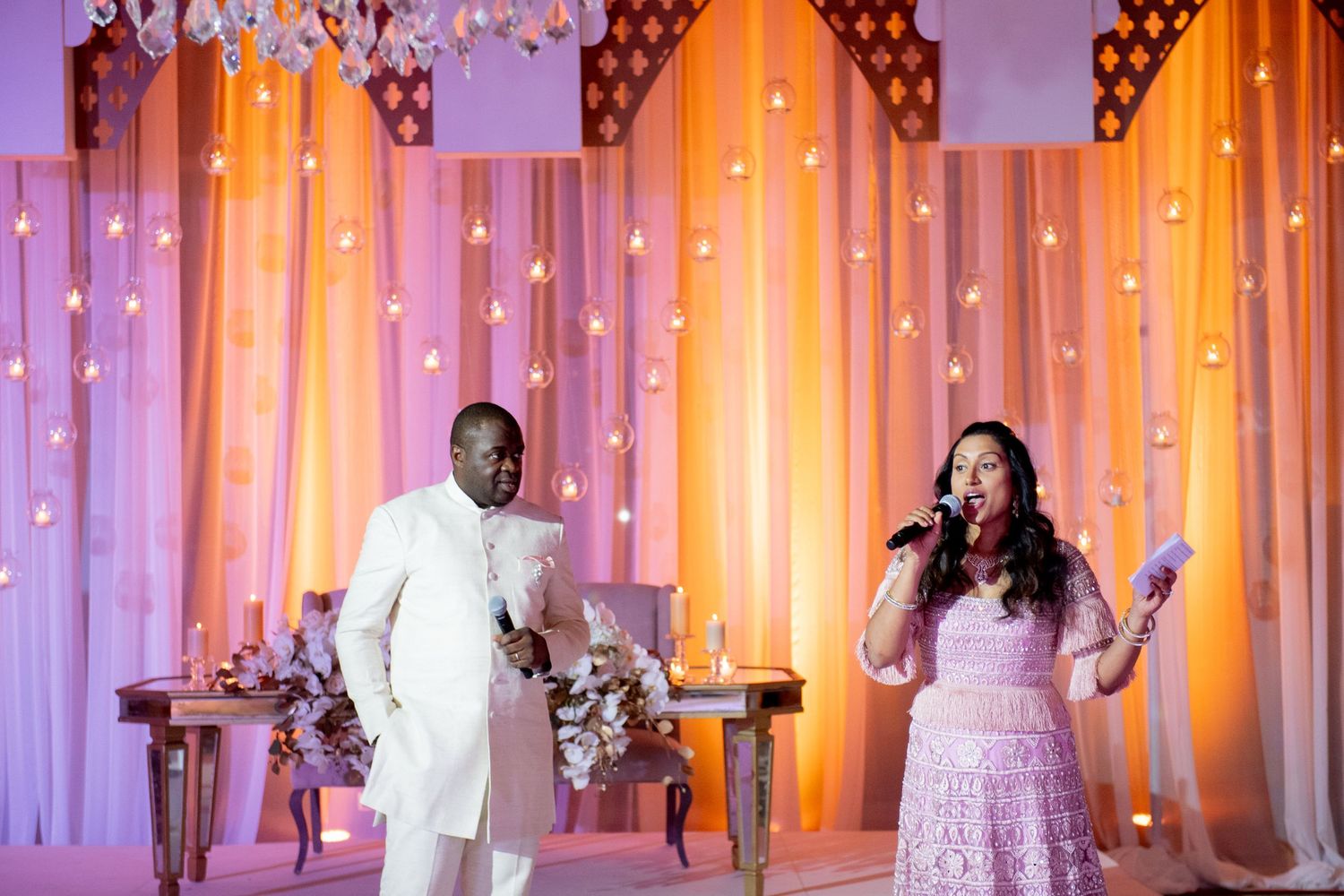 A man in a white suit and woman in pink lace dress perform at an elegant wedding reception with orange drape backdrop.