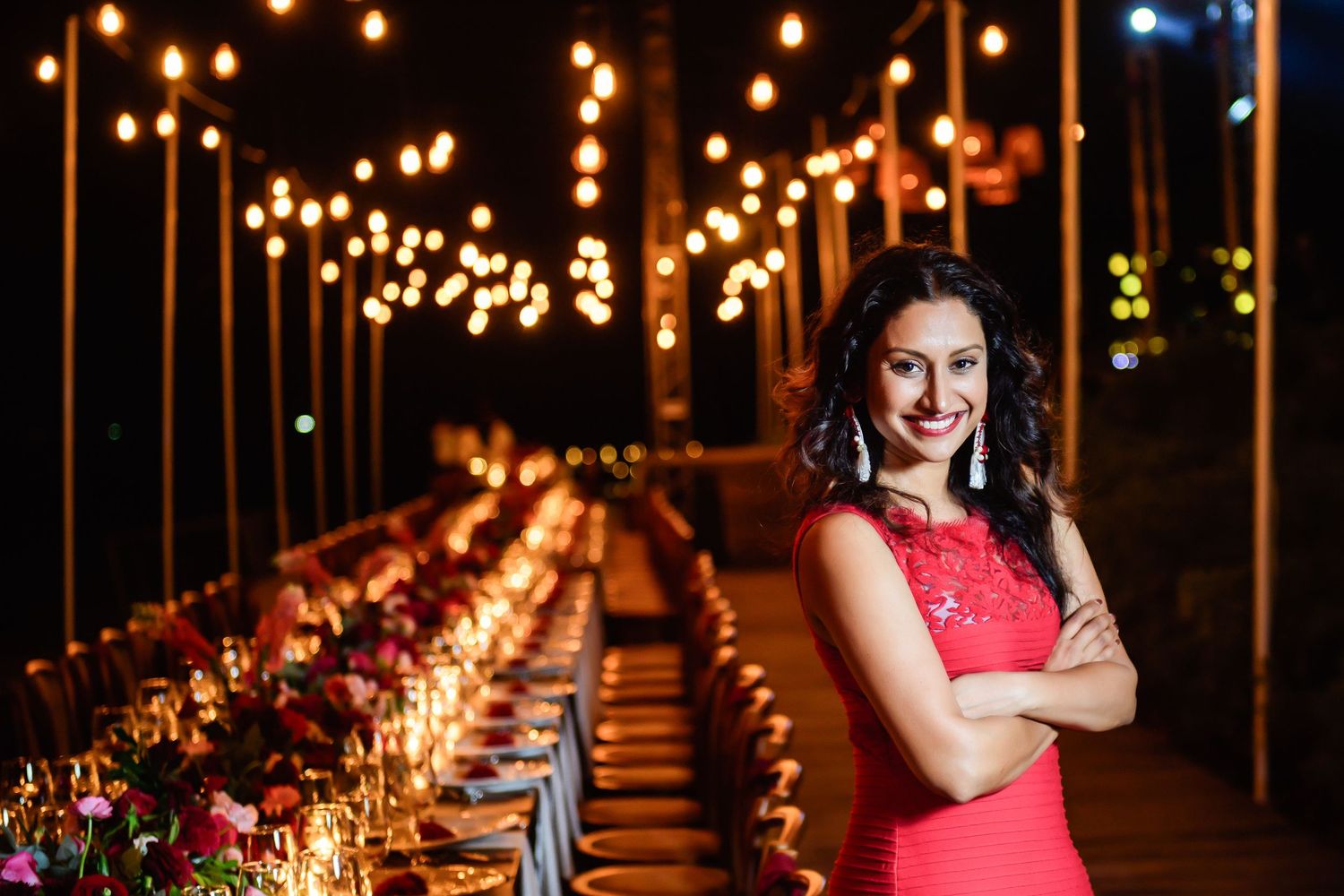 Woman in red dress smiling with arms crossed at elegantly lit outdoor dinner party with candles and string lights.