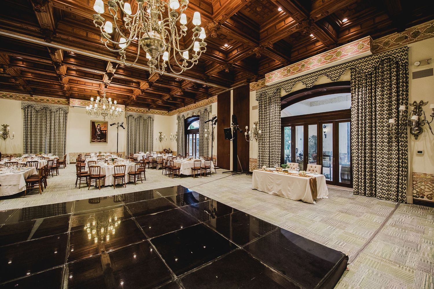 Elegant ballroom with black dance floor, chandelier, white-draped dining tables, and ornate wood-paneled ceiling.