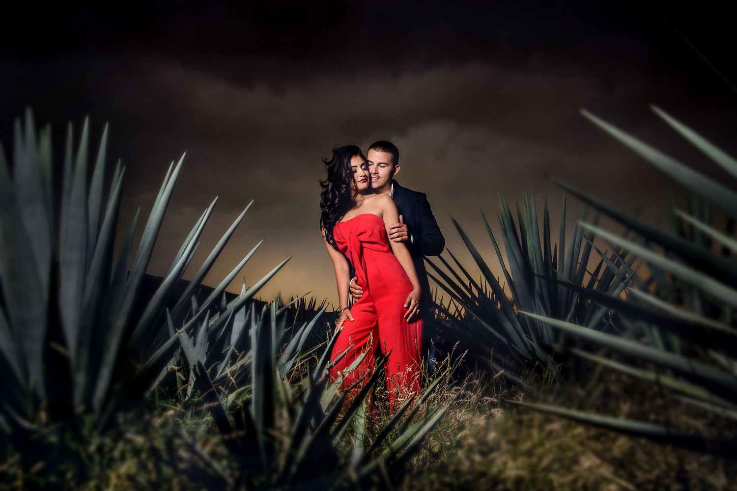 Couple embracing romantically in an agave field at night, woman in red jumpsuit and man in dark suit.