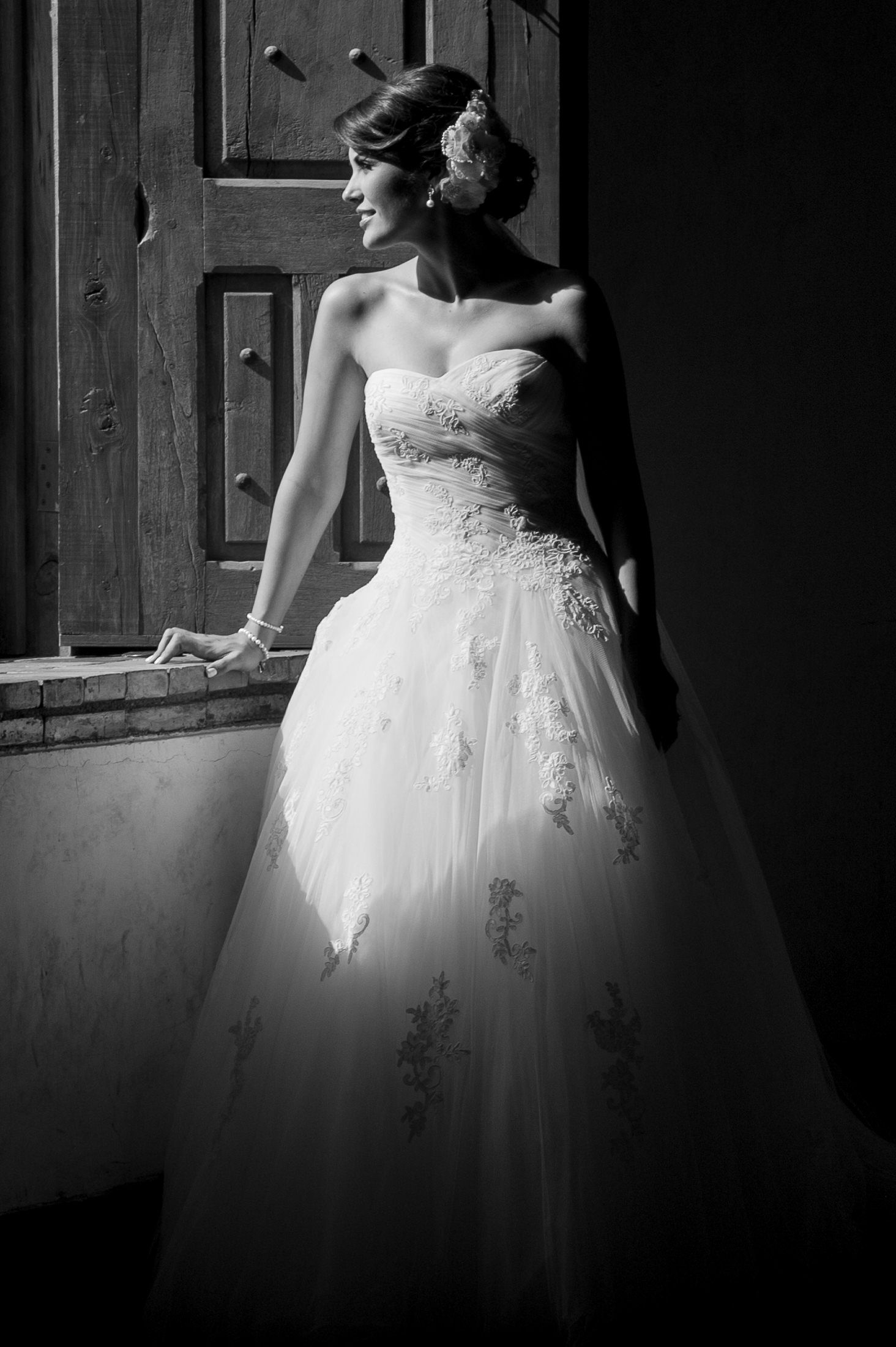 Dramatic black and white portrait of a bride in a strapless lace wedding gown looking to the side near a wooden door.
