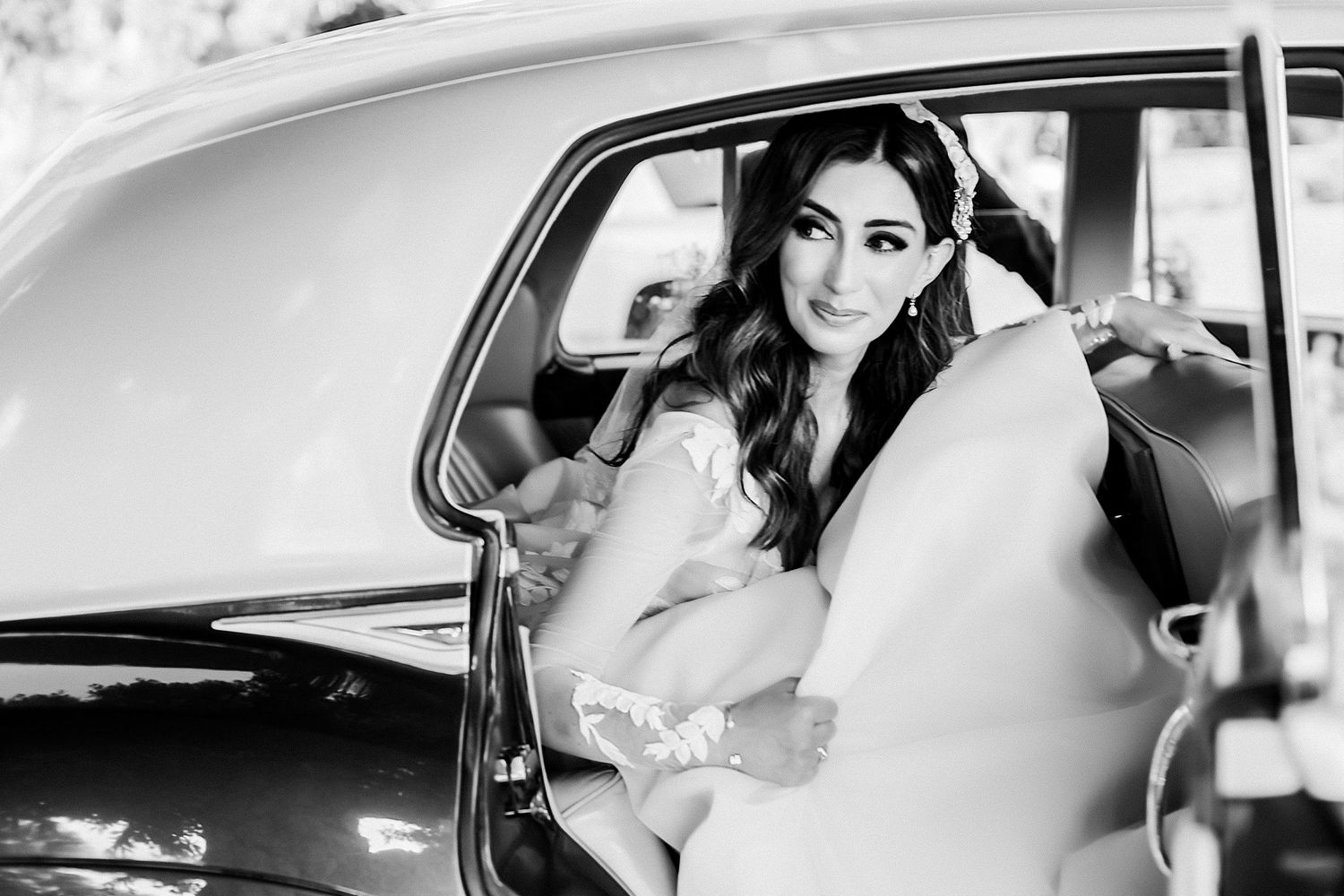 Black and white photo of a bride in an elegant dress sitting in a vintage car on her wedding day.