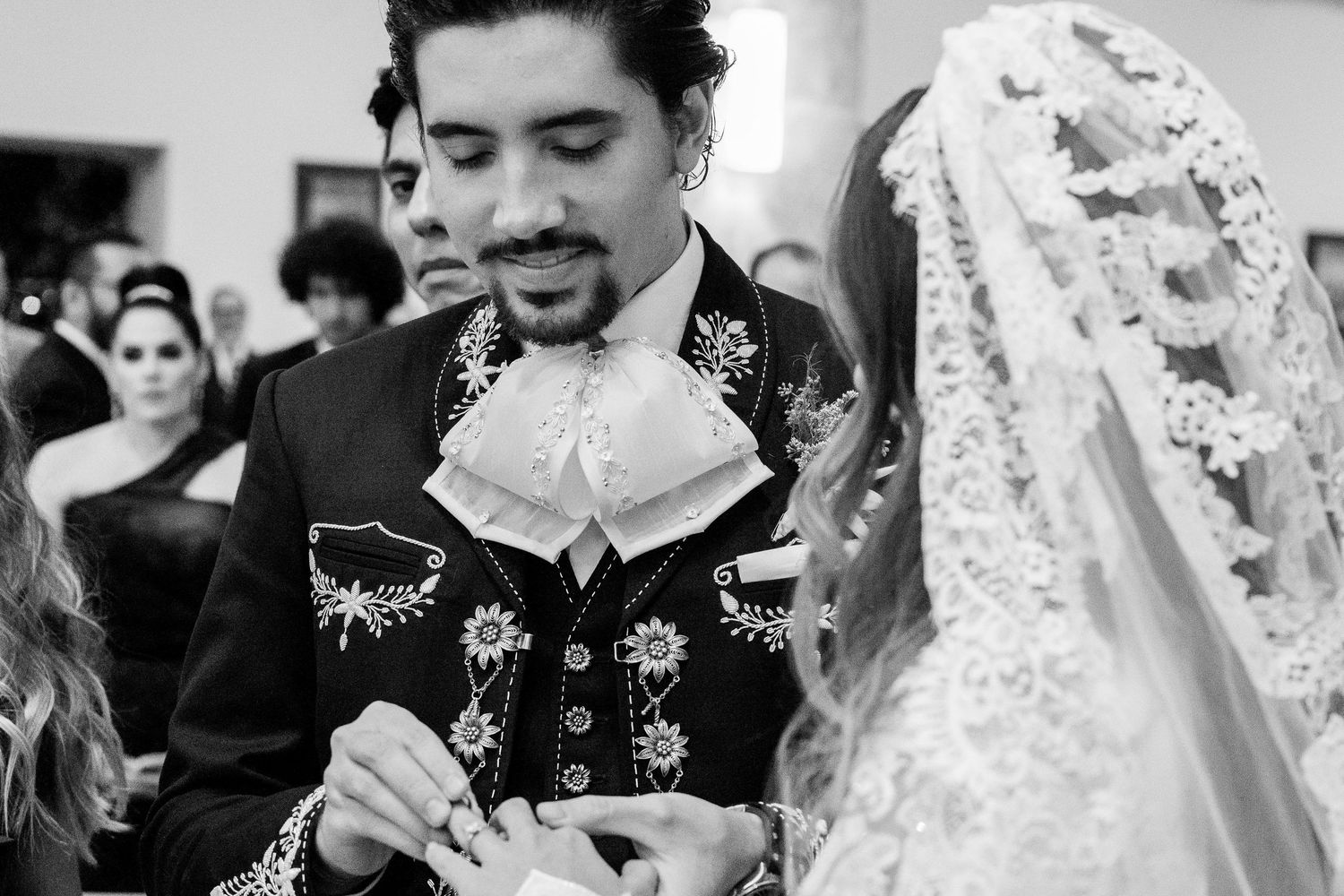 Groom in embroidered mariachi suit places ring on brides finger during wedding ceremony.